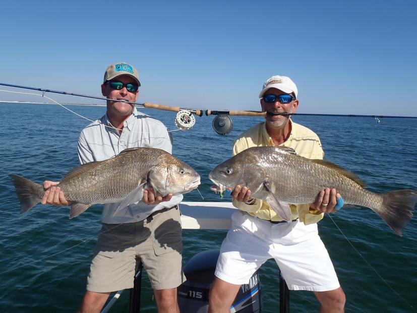 Two anglers, each with a black drum and a fly rod clamped in his teeth